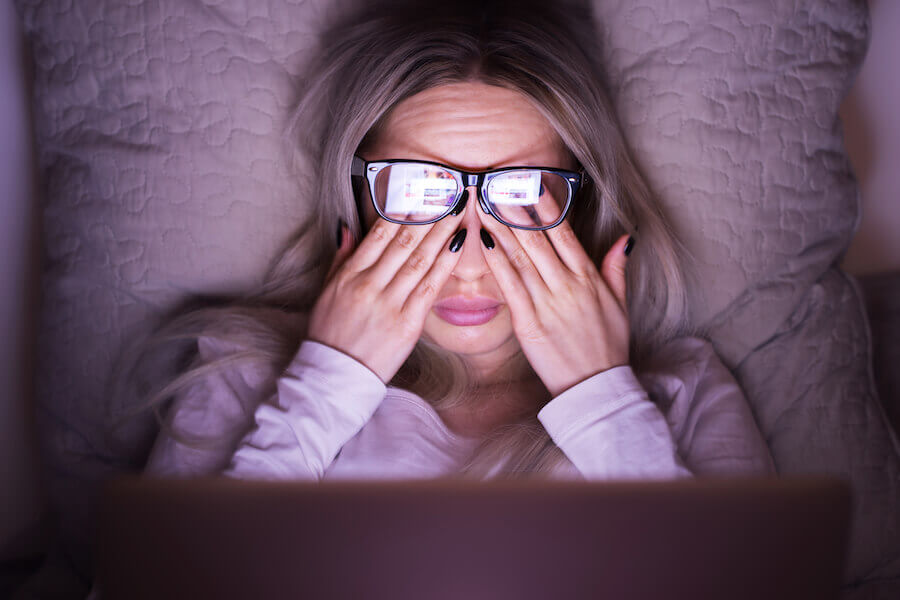 Woman rubbing eyes while laying back on a pillow with reflection of laptop in the glasses